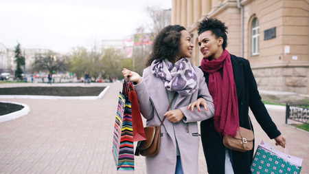 Two attractive mixed race women with shopping bags talking and walking down the street. Girlfriends have fun after visiting mall saleの写真素材