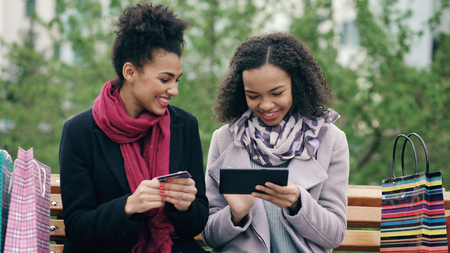 Two attractive mixed race women with paper bags sitting on bench and using tablet computer and credit card for online shoppingの写真素材