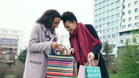 Two young african american women sharing their new purchases in shoppping bags with each other. Attractive girls talking and smiling after visiting mall salesの写真素材