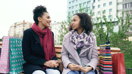 Two young african american women sharing their new purchases in shoppping bags with each other. Attractive girls talking while sitting on steet benchの写真素材