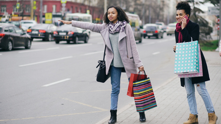 Two attractive african american women with shopping bags calling for taxi cab while coming back from mall salesの写真素材