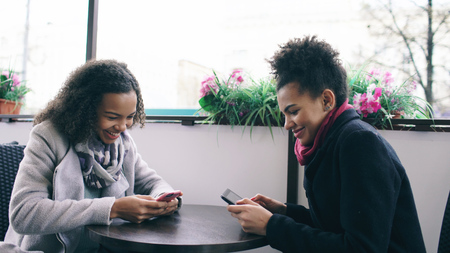 Two attractive mixed race female friends sharing together using smartphone in street cafe outdoorsの写真素材
