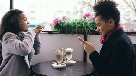 Two attractive mixed race female friends photographing coffee cups together using smartphone camera while sitting in street cafe outdoorsの写真素材
