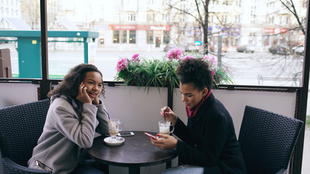 Attrcative mixed race woman sitting at the table in the street cafe talking cell phone while her friend surfing smartphoneの写真素材