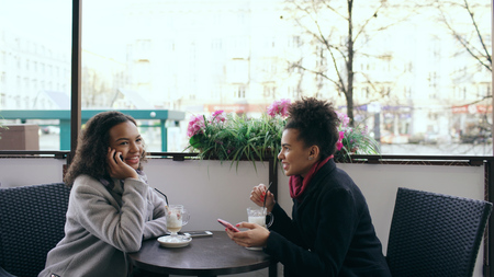 Attrcative mixed race woman sitting at the table in the street cafe talking cell phone while her friend surfing smartphoneの写真素材