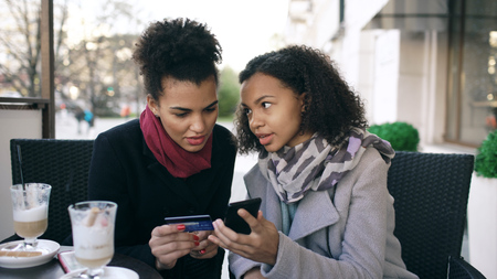 Two attractive mixed race women having online shopping with credit card and smartphone while talking and drinking coffee in street cafeの写真素材