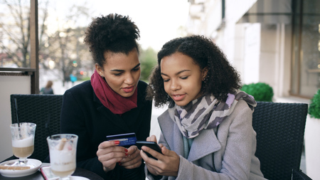 Two attractive mixed race women having online shopping with credit card and smartphone while talking and drinking coffee in street cafeの写真素材
