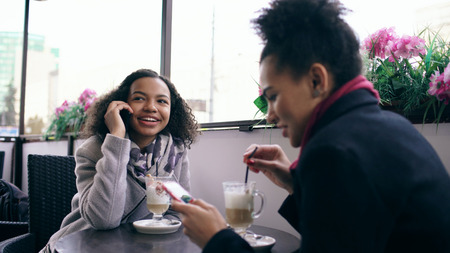 Two mixed race women sitting at the table in the street cafe using smartphone and talkingの写真素材