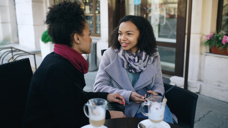 Two attractive mixed race women talking and drinking coffee in street cafe. Friends have fun after visiting mall saleの写真素材