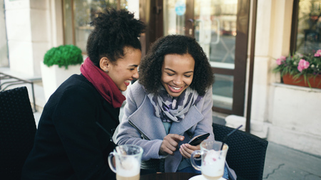 Two attractive mixed race female friends sharing together using smartphone in street cafe outdoorsの写真素材