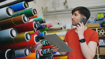 Young worker in warehouse talking smartphone and checking inventory. Man works in sales department of advertising materialsの写真素材