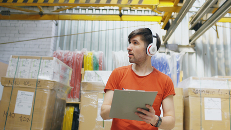 Happy young worker in industrial warehouse listening to music during work. Man in headphones have fun at workplace.の写真素材