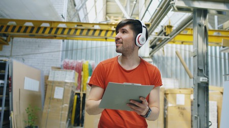 Happy young worker in industrial warehouse listening to music during work. Man in headphones have fun at workplace.の写真素材