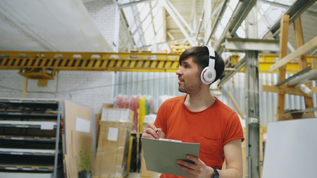 Happy young worker in industrial warehouse listening to music and dancing during work. Man in headphones have fun at workplace.の写真素材