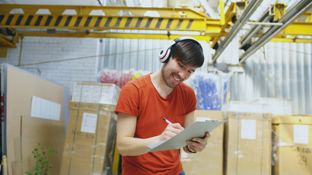 Happy young worker in industrial warehouse listening to music and dancing during work. Man in headphones have fun at workplace.の写真素材
