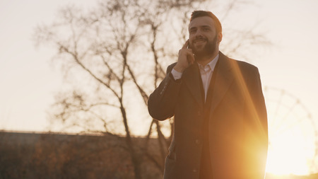 Young bearded business man in coat talking at smartphone and making deals on street with sunlight backlit in evening timeの写真素材