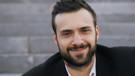 Close up portrait of bearded young businessman smiling and looking into camera in street outdoorsの写真素材