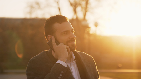Closeup of Young bearded business man in coat talking at smartphone and making deals on street with sunlight backlit in evening timeの写真素材