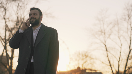 Young bearded business man in coat talking at smartphone and making deals on street with sunlight backlit in evening timeの写真素材