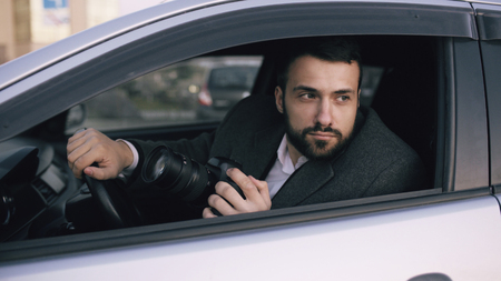 Young private detective man sitting inside car and photographing with dslr cameraの写真素材
