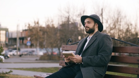 Creative young writer man in hat and coat write notes for his future book with pen in notebook sitting on city street bench at parkの写真素材