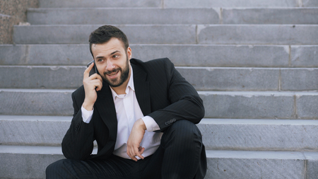 Young bearded business man talking on cell phone making deals sitting on stairs in streetの写真素材