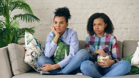 Two mixed race curly girl friends sitting on the couch and watch thriller movie and eat popcorn at homeの写真素材