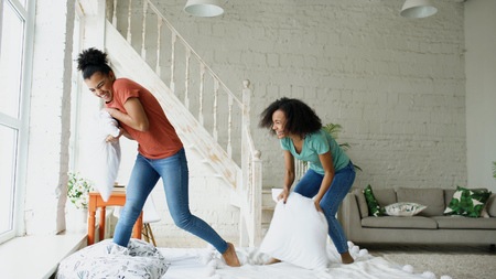 Mixed race young pretty girls jumping on bed and fight pillows having fun at homeの写真素材