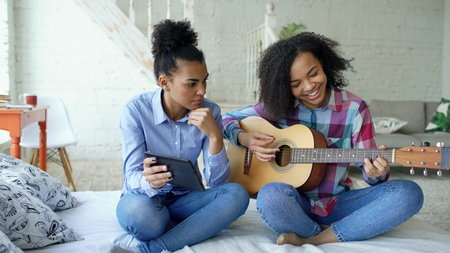 Mixed race young woman with tablet computer sitting on bed teaching her teenage sister to play acoustic guitar at homeの写真素材