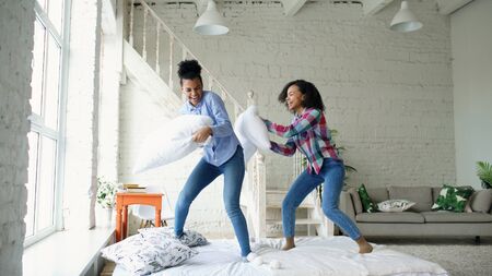 Mixed race young pretty girls jumping on bed and fight pillows having fun at homeの写真素材