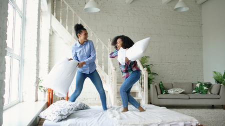 Mixed race young pretty girls jumping on bed and fight pillows having fun at homeの写真素材