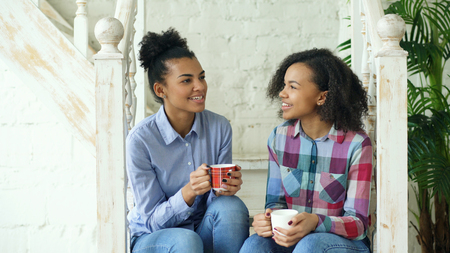 Two african american curly girls sistres sitting on stairs have fun laughing and chatting together at homeの写真素材