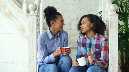 Two african american curly girls sistres sitting on stairs have fun laughing and chatting together at homeの写真素材
