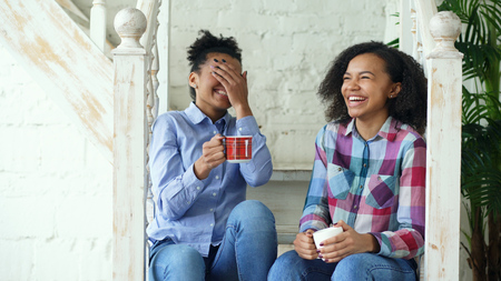 Two african american curly girls sistres sitting on stairs have fun laughing and chatting together at homeの写真素材