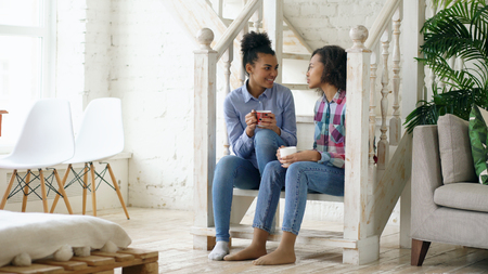 Two african american curly girls sistres sitting on stairs have fun laughing and chatting together at homeの写真素材