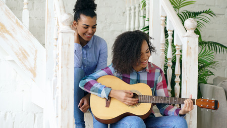Two brazilian curly girls sistres sitting on stairs and practice to play acoustic guitar. Friends have fun and singing at homeの写真素材