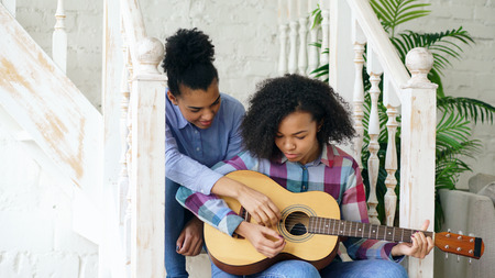 Mixed race young woman sitting on stairs teaching her teenage sister to play acoustic guitar at homeの写真素材