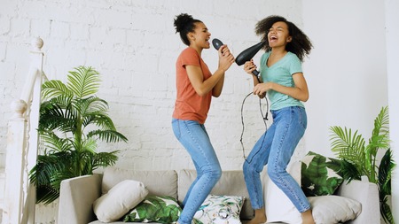 Mixed race young funny girls dance singing with hairdryer and comb jumping on sofa. Sisters having fun leisure in living room at home conceptの写真素材