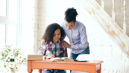 Teenage curly haired mixed race young girl sitting at the table concentrating focused learning lessons and her elder sister helps her studyingの写真素材