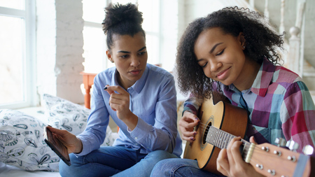 Mixed race young woman with tablet computer sitting on bed teaching her teenage sister to play acoustic guitar at homeの写真素材