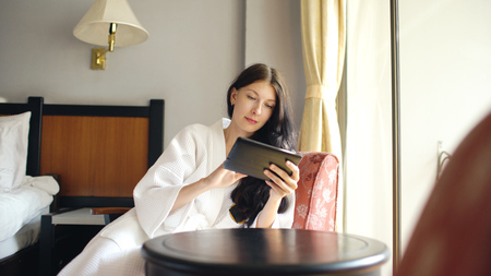 Pretty young woman in bathrobe chatting on tablet computer sitting on chair in hotel roomの写真素材