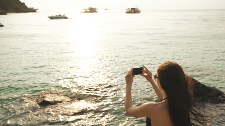 Woman tourist on beach island taking photograph of sunset with smartphone on holiday of boat and skyline viewの写真素材