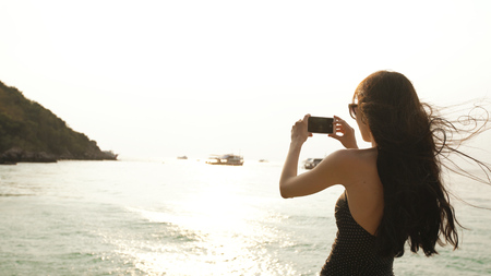 Woman tourist on beach island taking photograph of sunset with smartphone on holiday of boat and skyline viewの写真素材