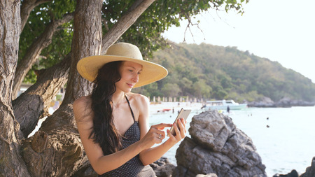 Young sexy woman in swimsuit and hat making selfie shoot using smartphone during vacation on beachの写真素材