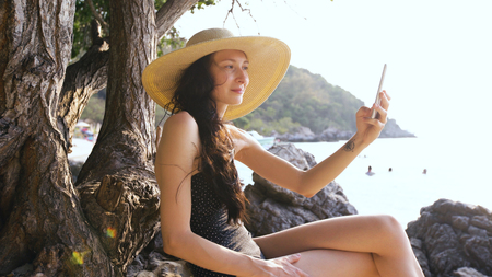 Young sexy woman in swimsuit and hat making selfie shoot using smartphone during vacation on beachの写真素材