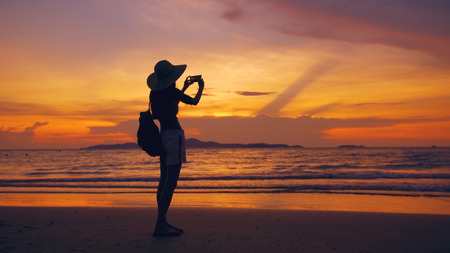 Silhouette of young tourist woman in hat taking photo with cellphone during sunset in ocean beachの写真素材