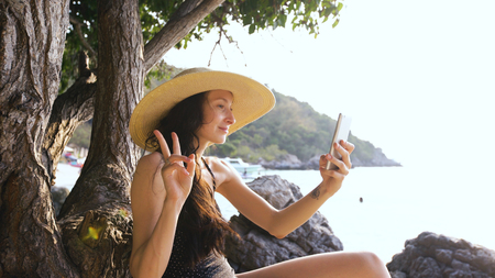 Young sexy woman in swimsuit and hat making selfie shoot using smartphone during vacation on beachの写真素材