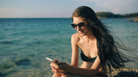 Beautiful woman sms texting using app on smart phone at beach sunset during vacationの写真素材