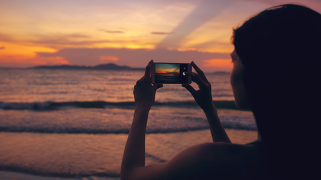 Closeup of young tourist woman photographs ocean view with smartphone during sunset at beachの写真素材
