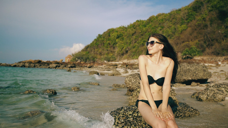 Young sexy woman in sunglasses smiling and sitting on rock at beautiful tropical beachの写真素材